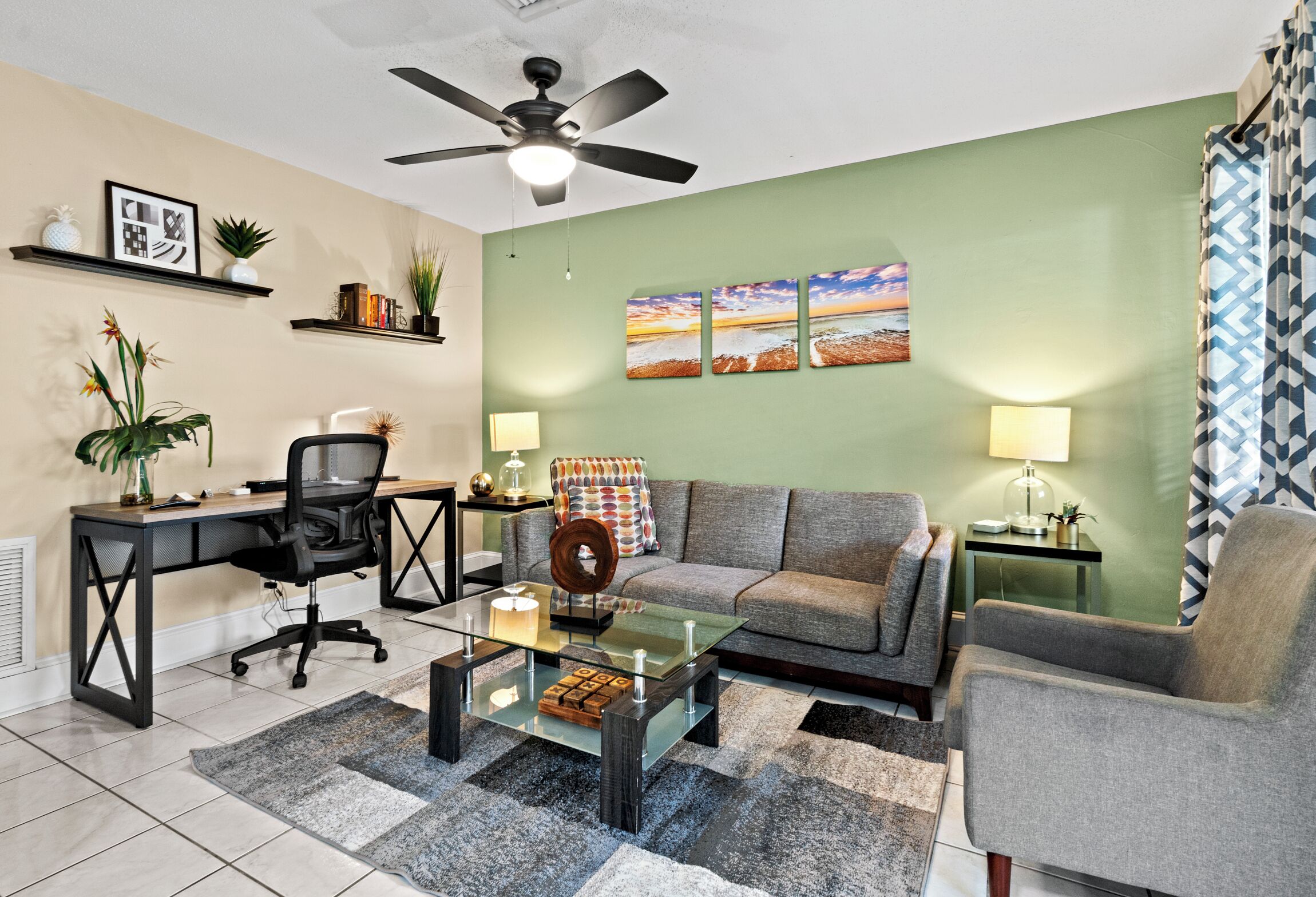 Un salon moderne avec une table basse en verre, un canapé gris, un fauteuil de bureau noir, un ventilateur de plafond et un mur d’accent vert.