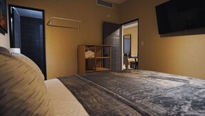 Un dormitorio con cama, una estantería de madera con toallas, un escritorio con silla y un televisor en la pared.