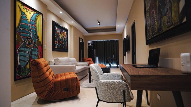 Una sala de estar moderna con un sofá, sillones, una mesa de centro y una computadora portátil sobre un escritorio de madera. Hay pinturas abstractas en las paredes y un comedor al fondo.