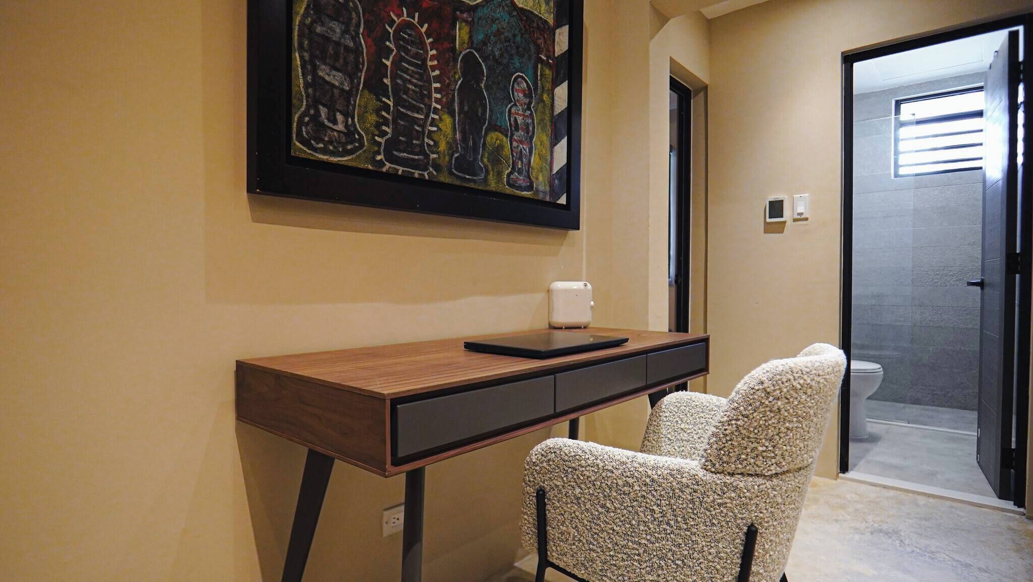 Une pièce comprenant un bureau en bois, une chaise, un tableau accroché au mur et une porte donnant sur une salle de bain.