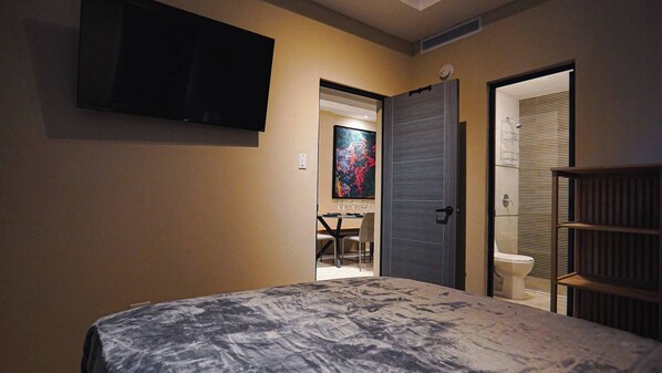 A bedroom with a bed, a TV mounted on the wall, a door leading to a bathroom, and a small table with chairs.