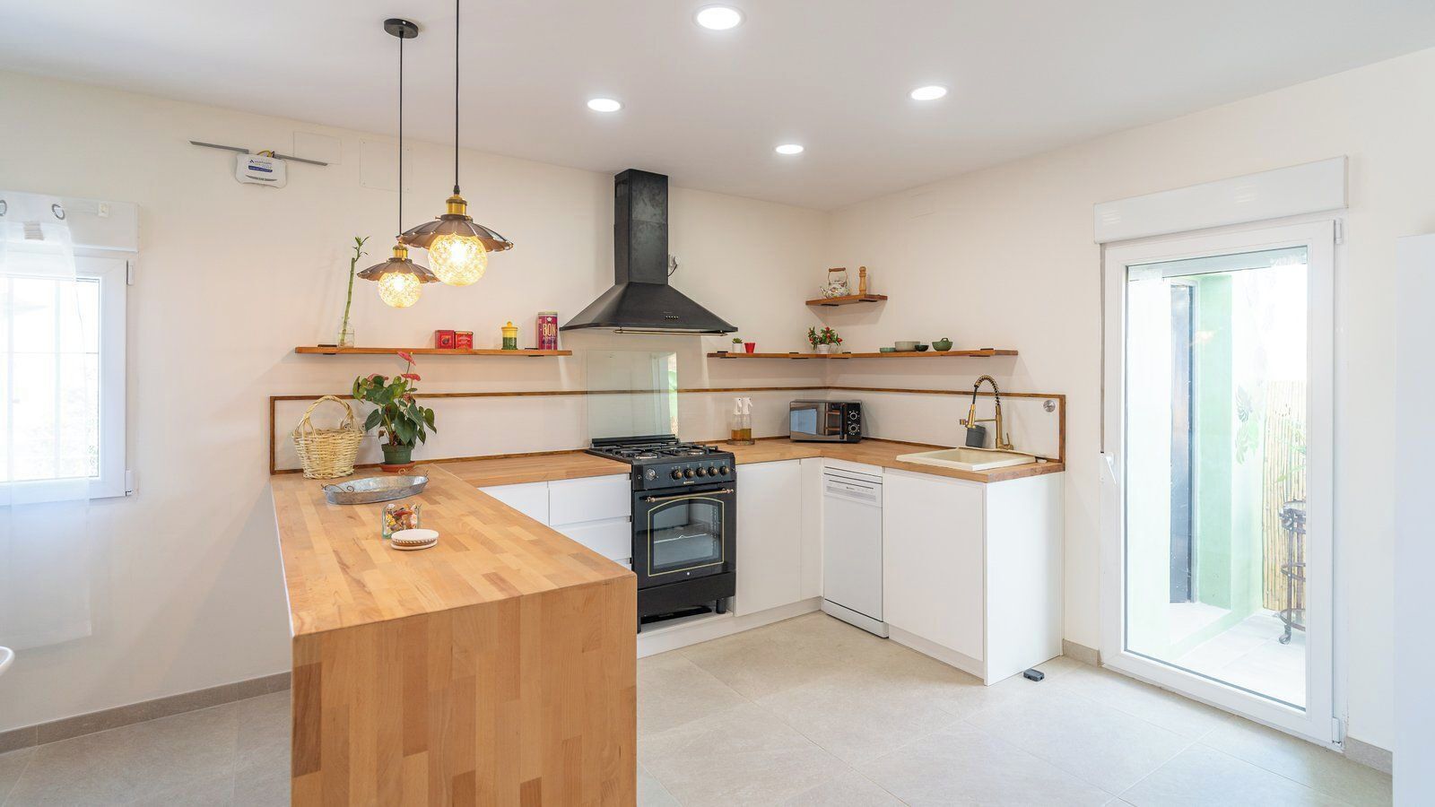 A modern kitchen with a central island, built-in oven, microwave, and dishwasher. There are pendant lights above the island and a black range hood above the stove.