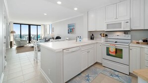 A modern kitchen with white cabinetry, a large island, a microwave, and an oven. There is a view of the city from the adjacent living area. - Dune View|Indoor Pool·Beachfront·Hot Tub·AB Condo (Atlantic Beach)