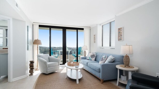 A modern living room with a sofa, armchair, coffee table, and a view of the city through large windows. - Dune View|Indoor Pool·Beachfront·Hot Tub·AB Condo (Atlantic Beach)