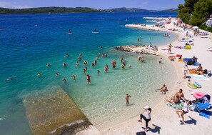 Ein Strand mit klarem Wasser, badenden Personen und einem sandigen Ufer mit Sonnenschirmen und Liegestühlen.