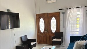 Een woonkamer met een grote flatscreen televisie aan de muur, een houten deur met glaspanelen, een blauwe bank, een witte salontafel en een rieten fauteuil.