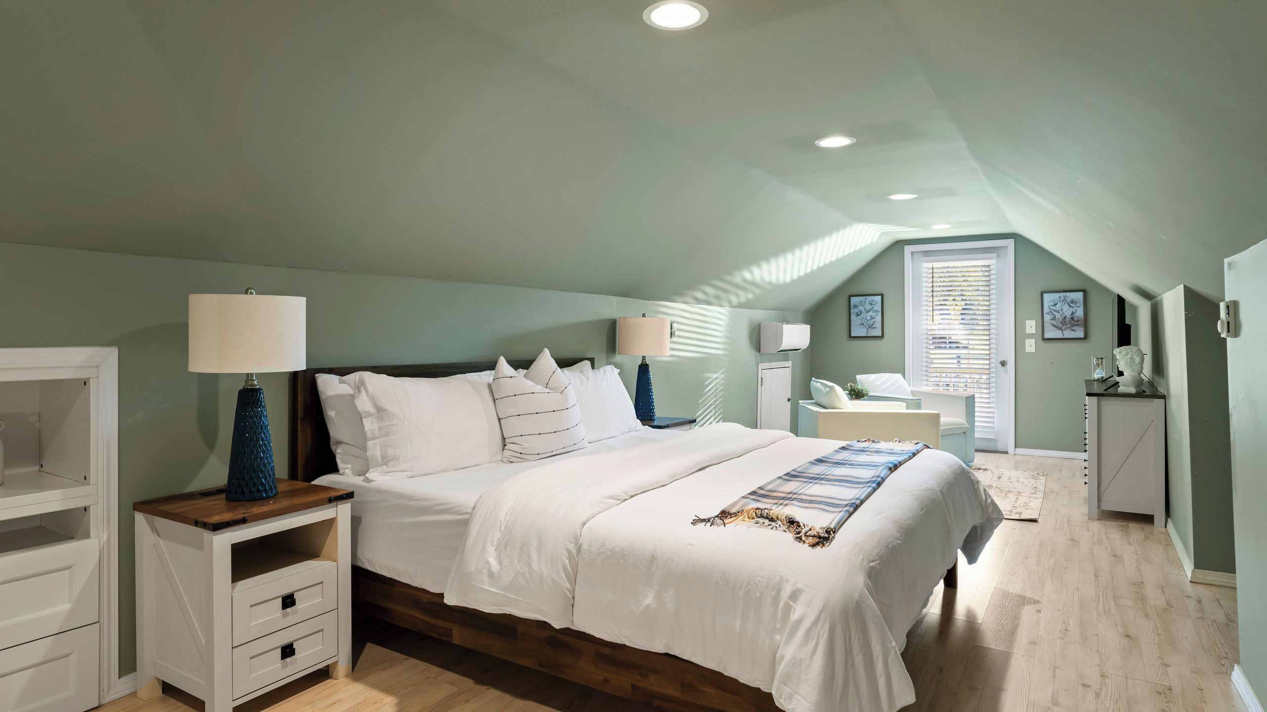 A bedroom with a sloped ceiling, a bed with white bedding, a nightstand with a lamp, a dresser, and a window with blinds.