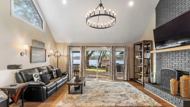 A modern living room with a leather sofa, a chandelier, a fireplace, and a view of the outdoors. - Lakefront Home w/ Game Room & Hot Tub Ponderosa Peninsula by Avantstay (Mooresville)