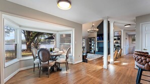 A spacious dining area with a large window overlooking a scenic view, a chandelier, and a cozy seating area with a leather chair. - Lakefront Home w/ Game Room & Hot Tub Ponderosa Peninsula by Avantstay (Mooresville)