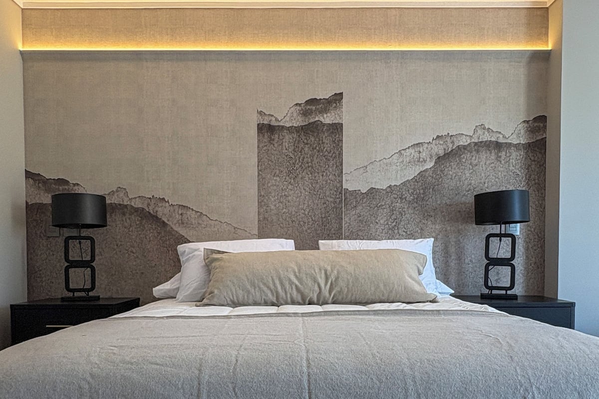 Un grand lit recouvert d’une couvre-lit à motifs, deux tables de chevet avec des lampes, et un mur orné d’une fresque représentant un paysage de montagne.