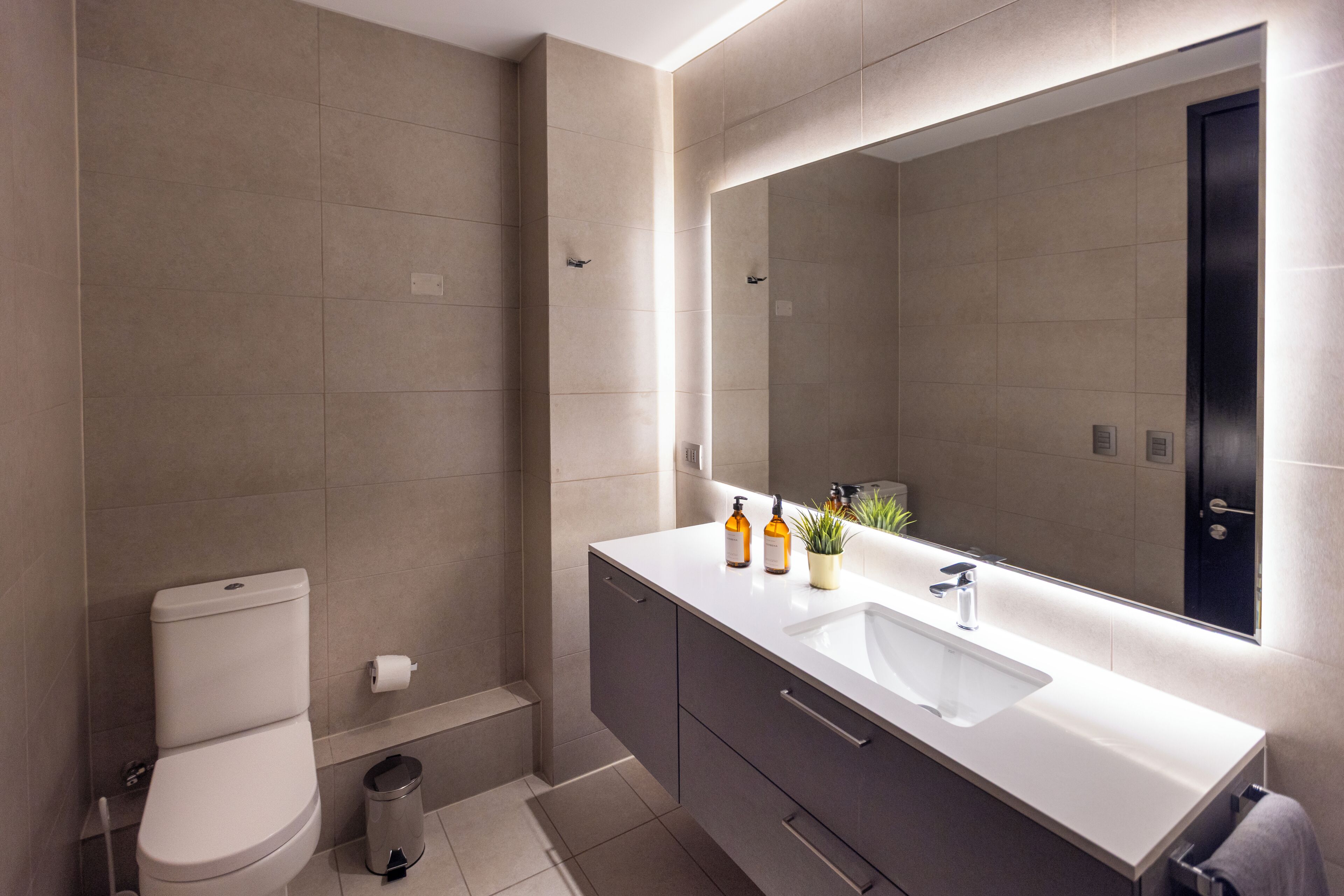 A modern bathroom with a toilet, sink, mirror, and a trash bin.
