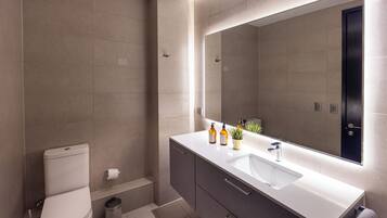 A modern bathroom with a toilet, sink, mirror, and a trash bin.
