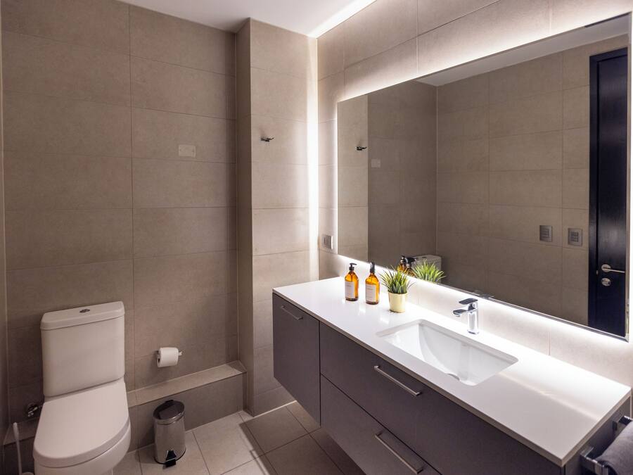A modern bathroom with a toilet, sink, mirror, and a trash bin.