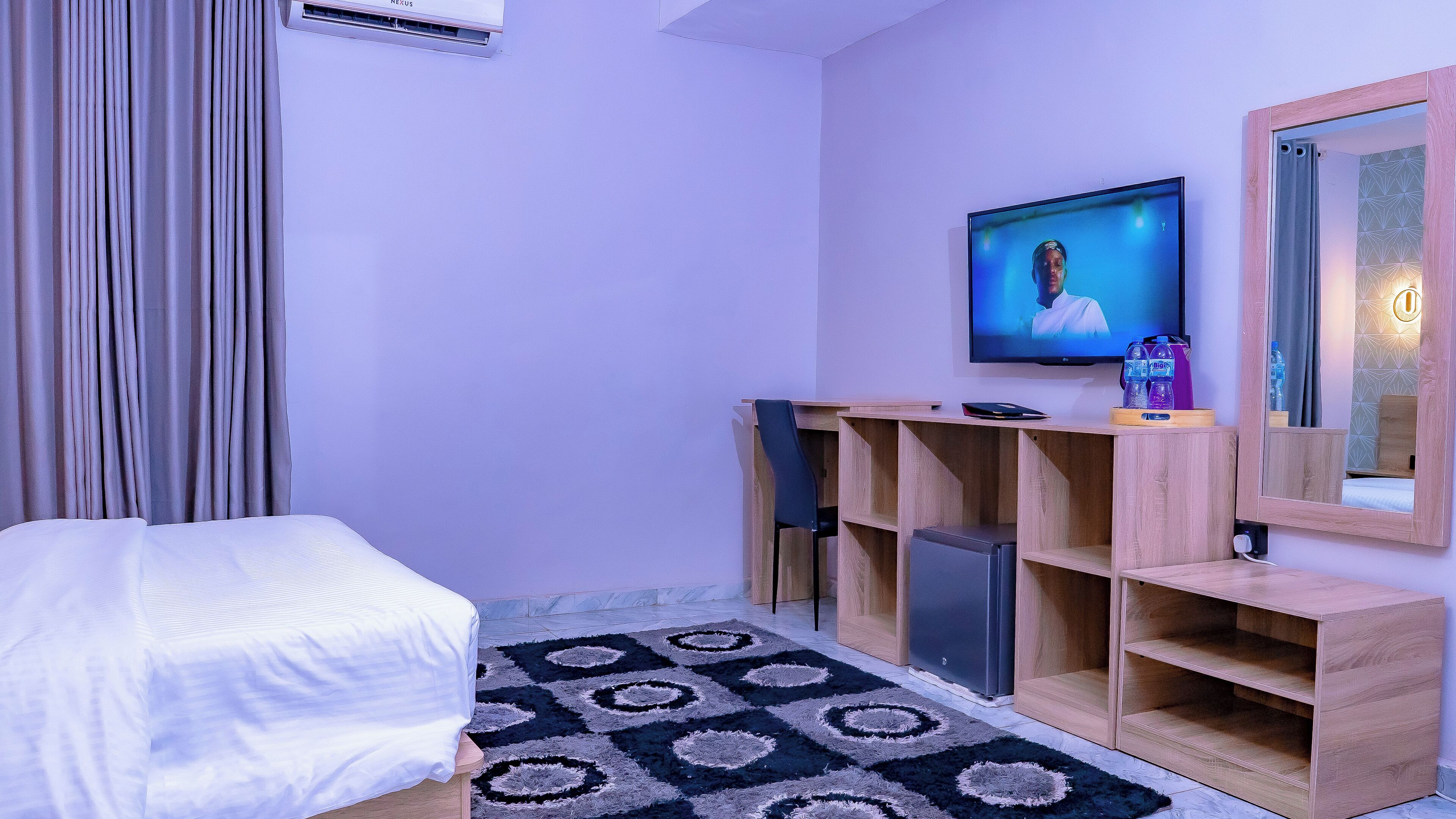 A hotel room with a bed, a TV, a wooden desk, a mirror, and a patterned rug.