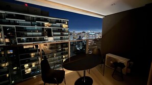 Un balcón con mesa y sillas, vista a un paisaje urbano al atardecer y un edificio moderno al fondo.