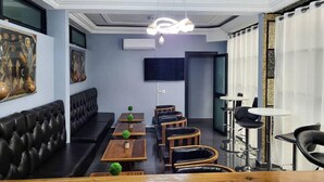 A modern lounge area with a black leather sofa, wooden tables, and chairs, a flat-screen TV, and a ceiling light fixture. - Justina Guest House Hotel  (Douala)