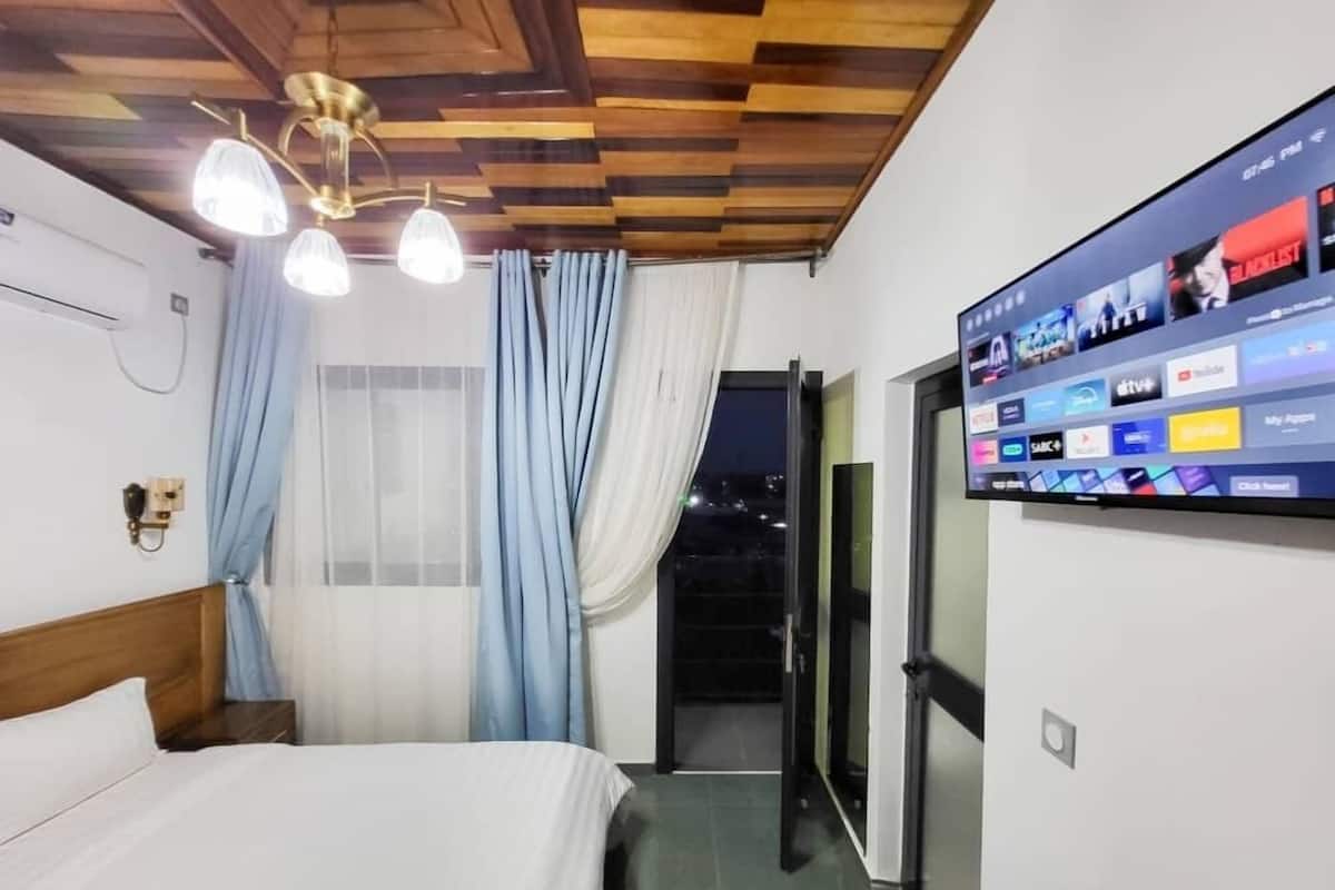 A hotel room with a wooden ceiling, a bed with white bedding, a wall-mounted TV displaying various channels, and a window with blue curtains.