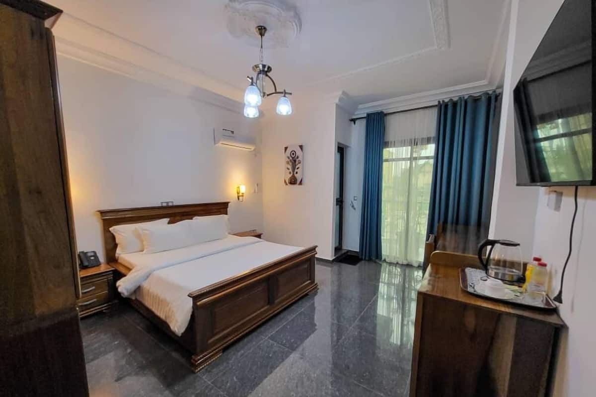 A hotel room with a large bed, a desk with a kettle, a television, and a window with curtains.