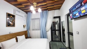 A hotel room with a wooden ceiling, a bed with white bedding, a mounted TV displaying various channels, and a window with blue curtains. - Justina Guest House Hotel  (Douala)
