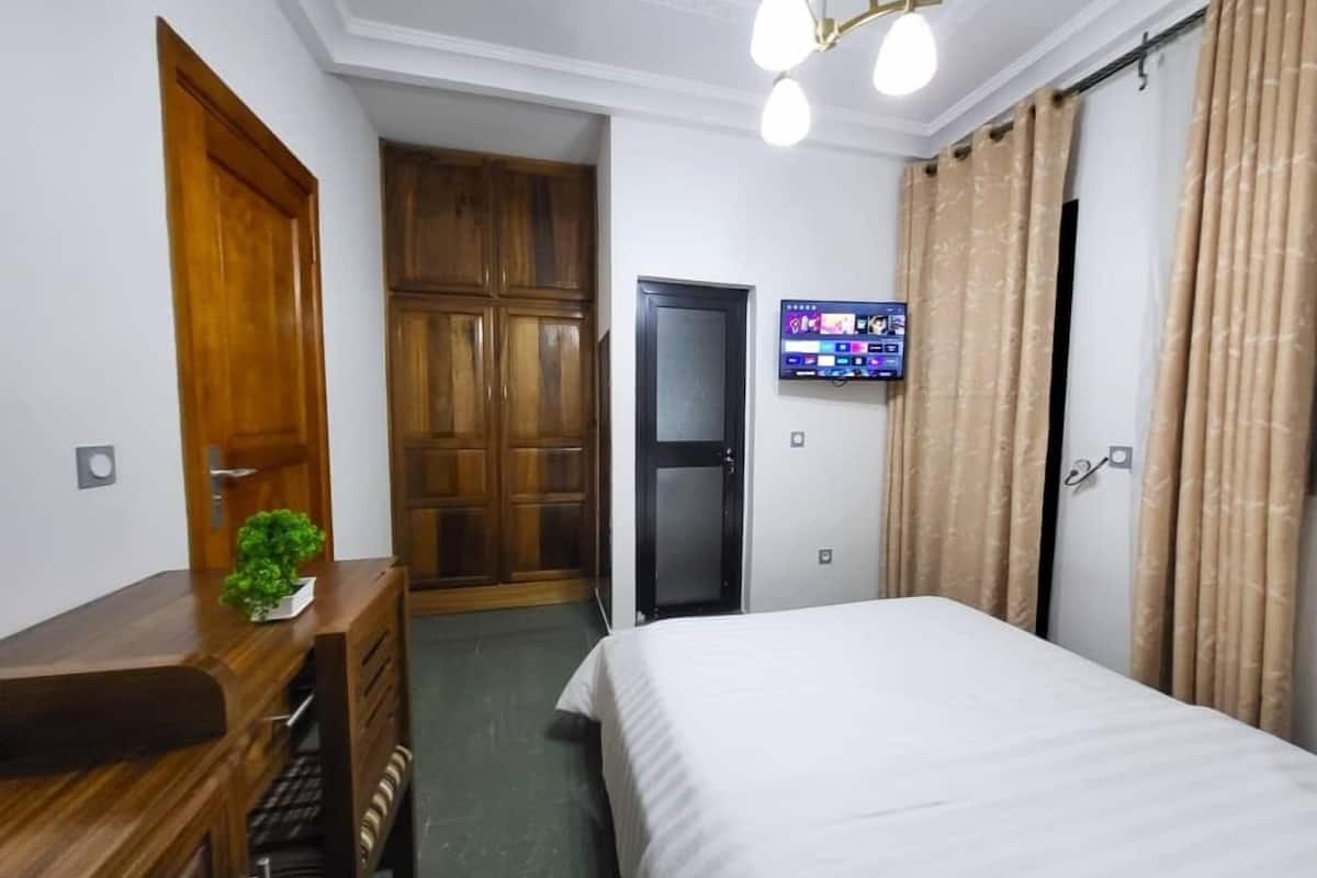 A hotel room with a wooden desk, a bed, a TV mounted on the wall, and a wardrobe.