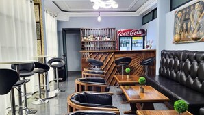 A modern bar area with wooden furniture, a Coca-Cola refrigerator, and a seating area with leather sofas and chairs. - Justina Guest House Hotel  (Douala)