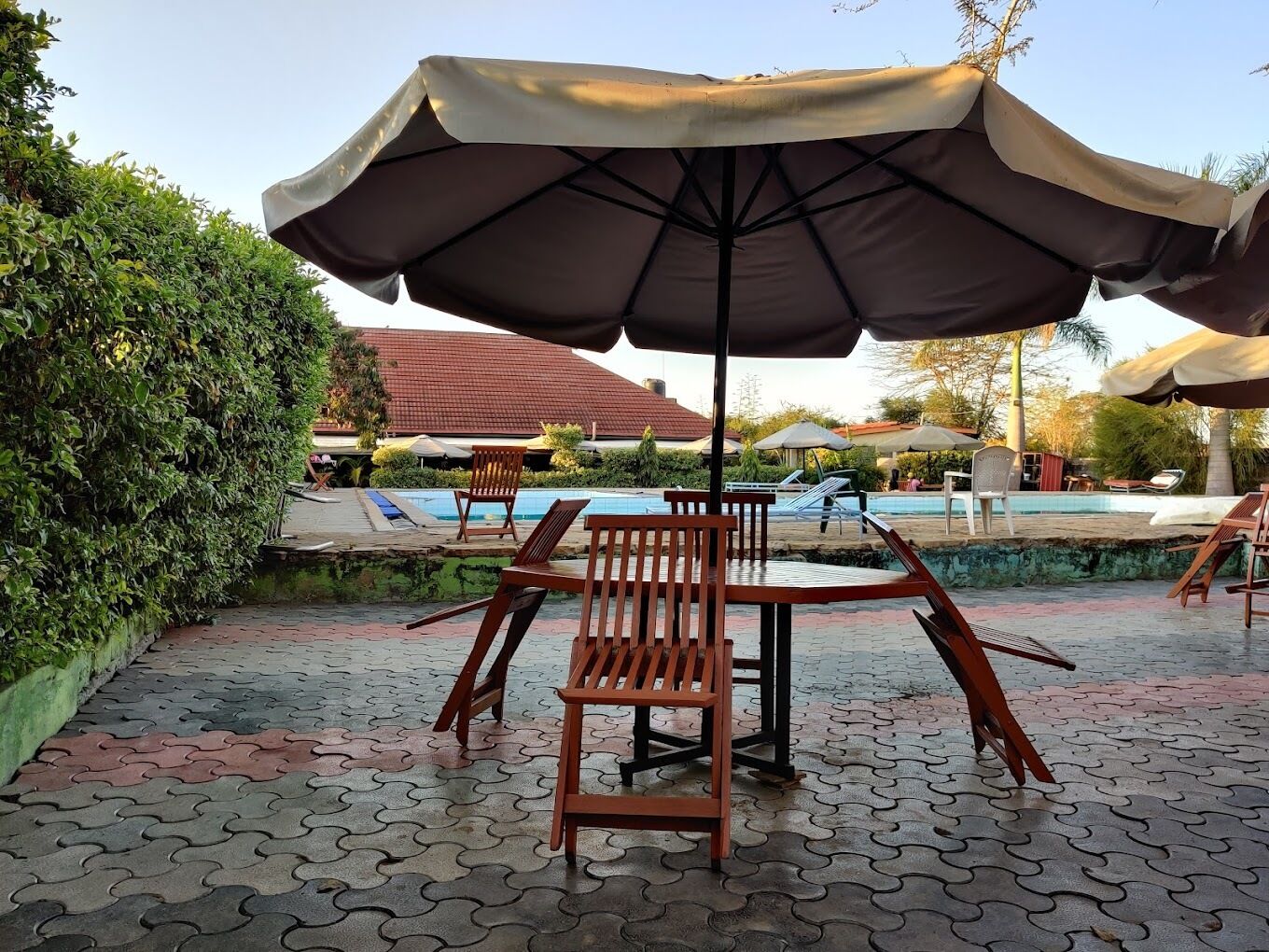 A poolside area with a table and chairs under a large umbrella, surrounded by a tiled patio and a swimming pool.