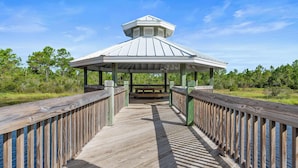 A wooden boardwalk leading to a gazebo with a peaked roof, surrounded by greenery.