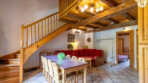 Una escalera de madera conduce a un comedor con una mesa y sillas, un sofá rojo, y un techo con vigas a la vista y una araña.