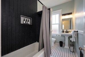 A bathroom with a black and white herringbone wall pattern, a white bathtub, a shower curtain, a sink with a mirror, and a tiled floor. - The Old Fashioned | Hot Tub, Fire Pit, Mini Golf Steps From Bardstown Rd (Louisville)