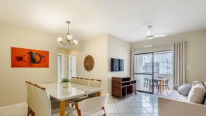 Una sala moderna con puerta de vidrio que da a un balcón, un comedor con mesa de vidrio y un televisor montado en la pared.