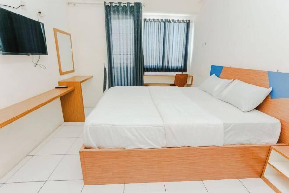 A bedroom with a bed, a TV, a desk, and a window with curtains.