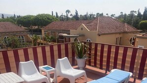 A rooftop terrace with white lounge chairs and a small table, surrounded by potted plants.