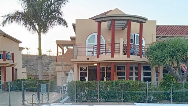 A swimming pool with a tiled deck, a palm tree, and a building with a balcony in the background.