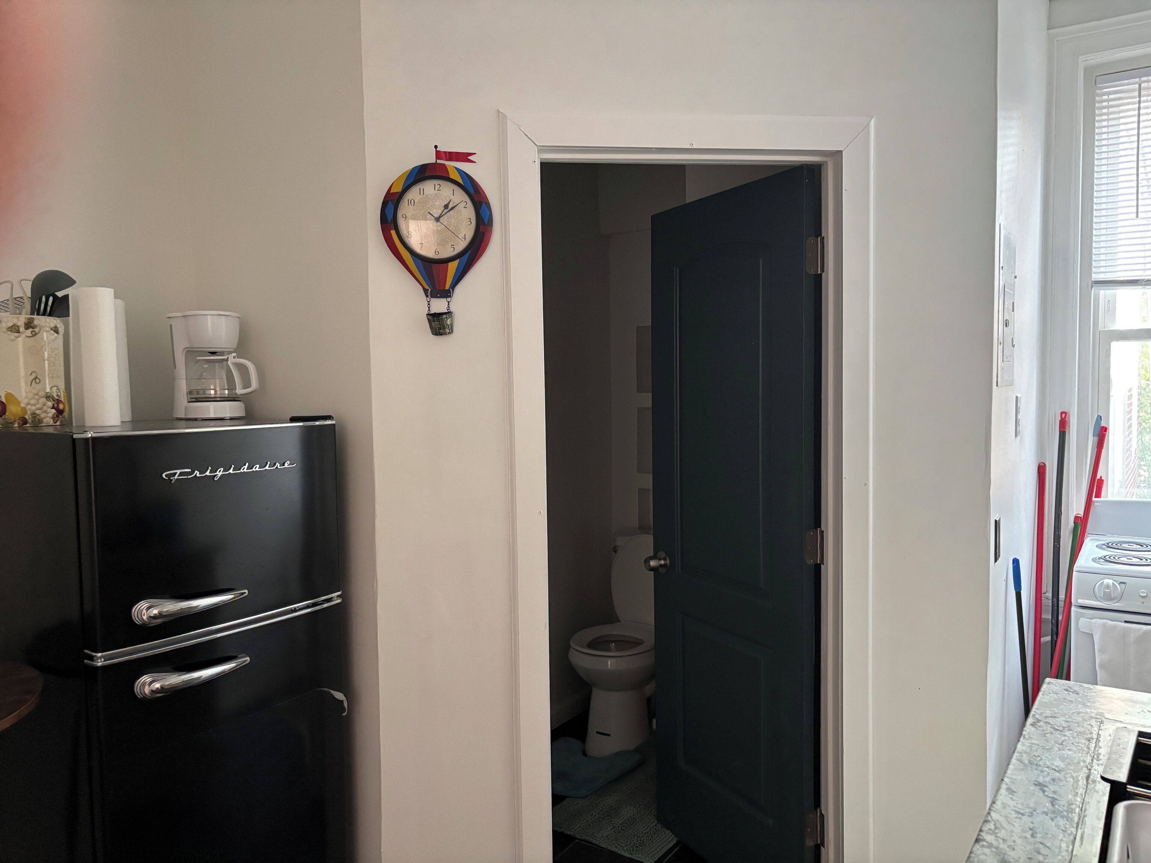 A compact kitchen with a black fridge, a coffee maker, and a colorful clock on the wall.