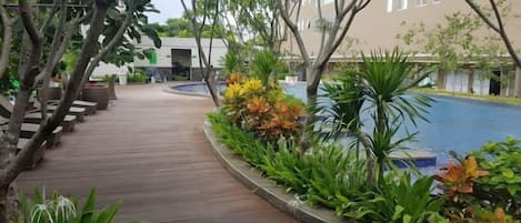 Uma passarela de madeira leva a uma área de piscina, com diversos tipos de plantas e um edifício ao fundo.