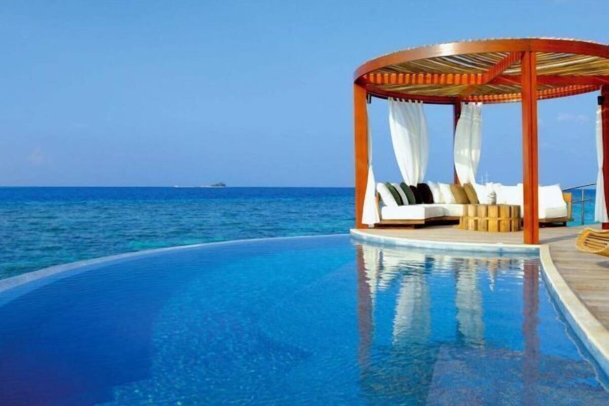 Une piscine à débordement circulaire, agrémentée d’un gazebo en bois et de coussins blancs.