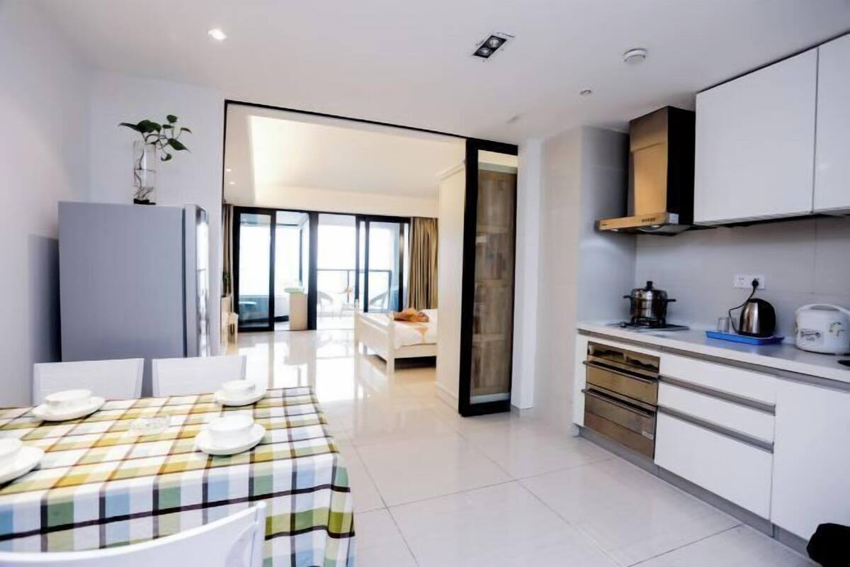 Une cuisine moderne avec des meubles blancs, une table à manger pour quatre personnes et une vue sur un espace de vie comprenant un canapé et une fenêtre.