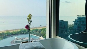 A modern bathroom with a bathtub, a window with a view of the sea and buildings, and a vase with a flower. - One Room One Car Seaview Holiday Apartment (Beihai)