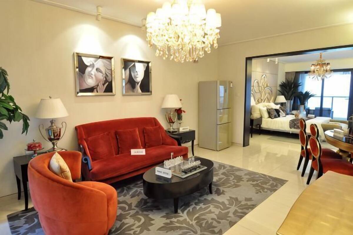 Un salon moderne avec un canapé rouge, une table basse et un lustre. Deux tableaux encadrés ornent le mur, et on aperçoit un coin repas avec une table et des chaises, ainsi qu’une cuisine au fond.