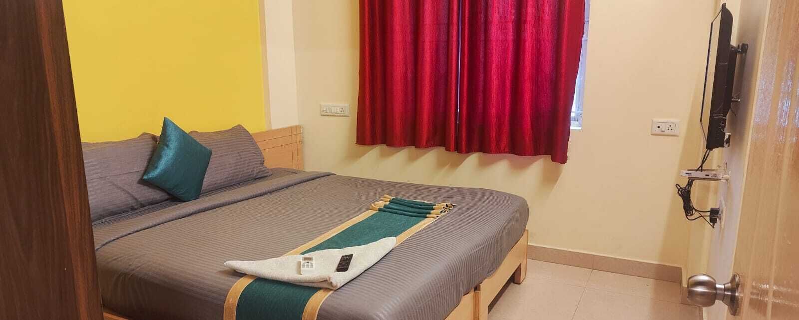A bedroom with a bed, pillows, a blanket, a window with red curtains, and a television mounted on the wall.
