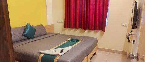 A bedroom with a bed, pillows, a blanket, a window with red curtains, and a television mounted on the wall.
