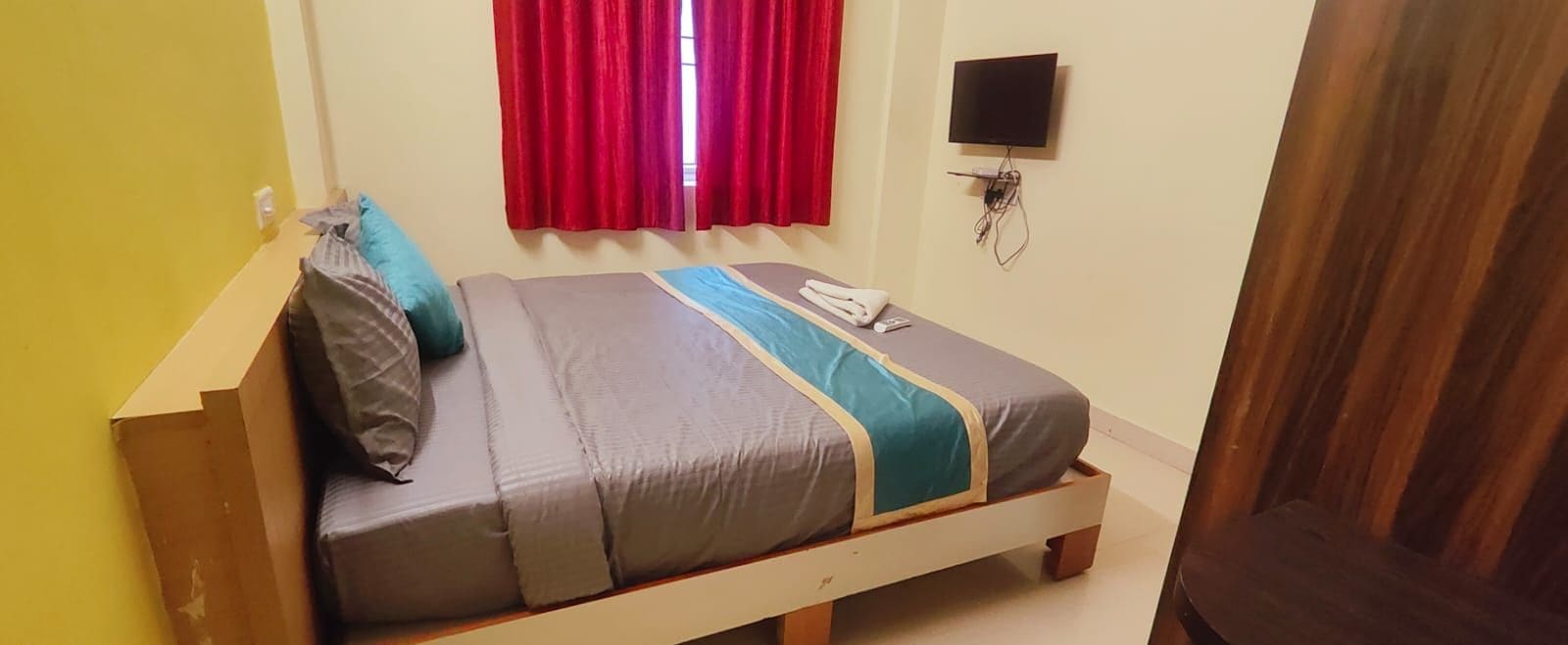 Una cama individual con estructura de madera, ropa de cama gris y un detalle en color verde azulado. Un televisor está montado en la pared sobre la cama.