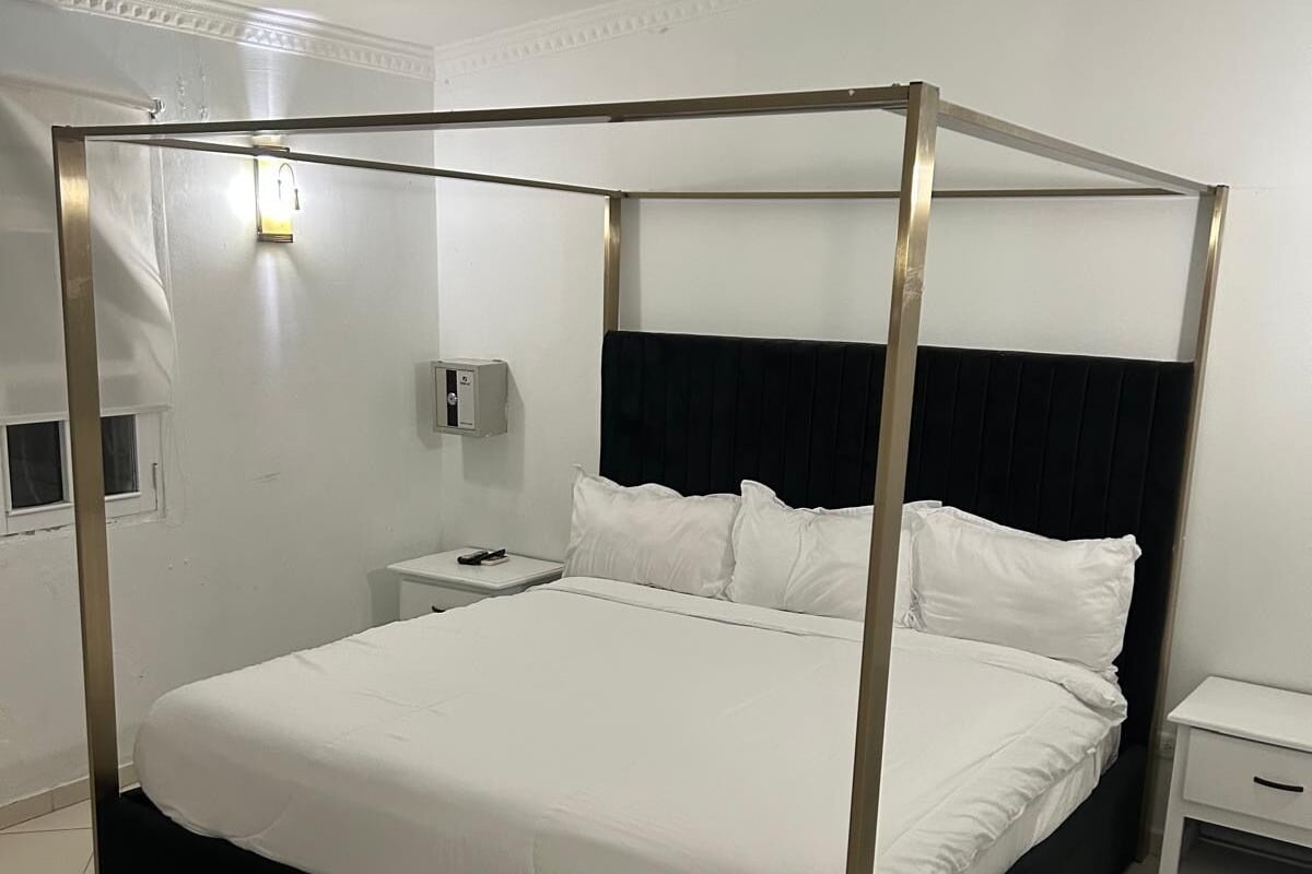 Een hemelbed met witte beddengoed, een zwart hoofdeinde en een nachtkastje met een lamp.