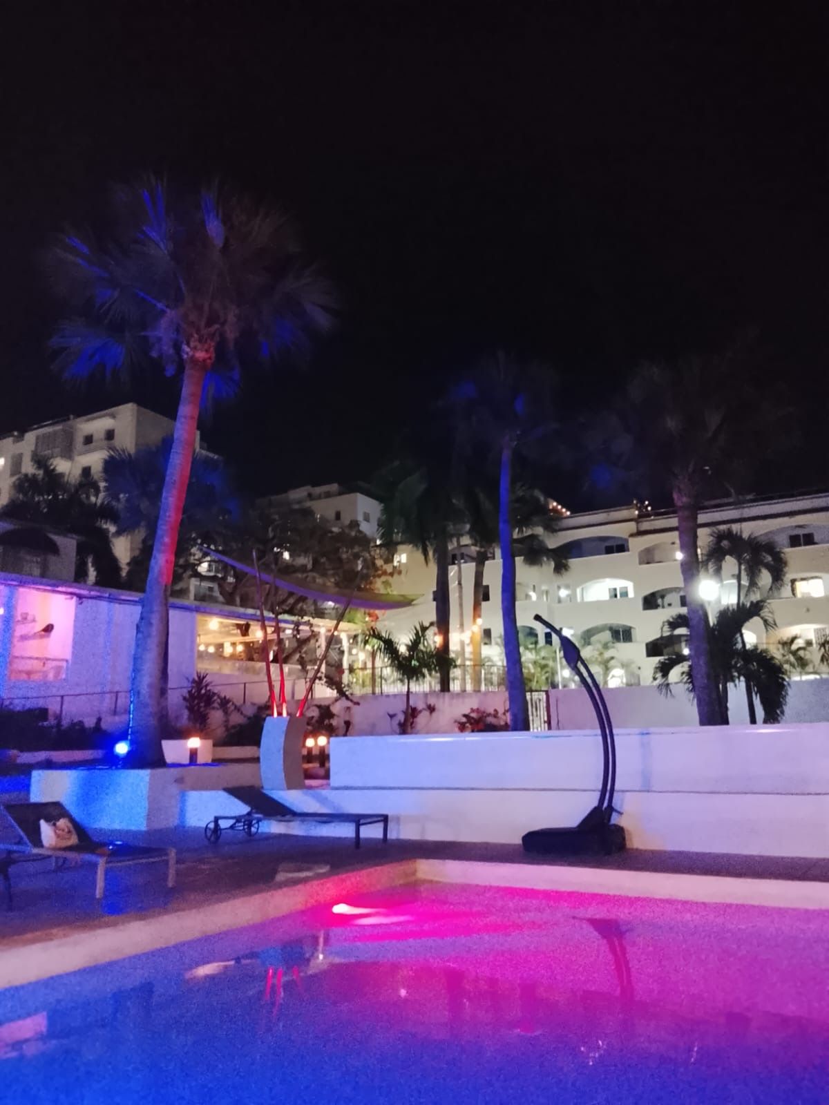 Vista nocturna de un área de piscina con palmeras y un edificio al fondo.