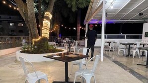 Un espace extérieur aménagé pour les repas, avec des tables et des chaises, un arbre décoré de guirlandes lumineuses et un coin salon abrité.