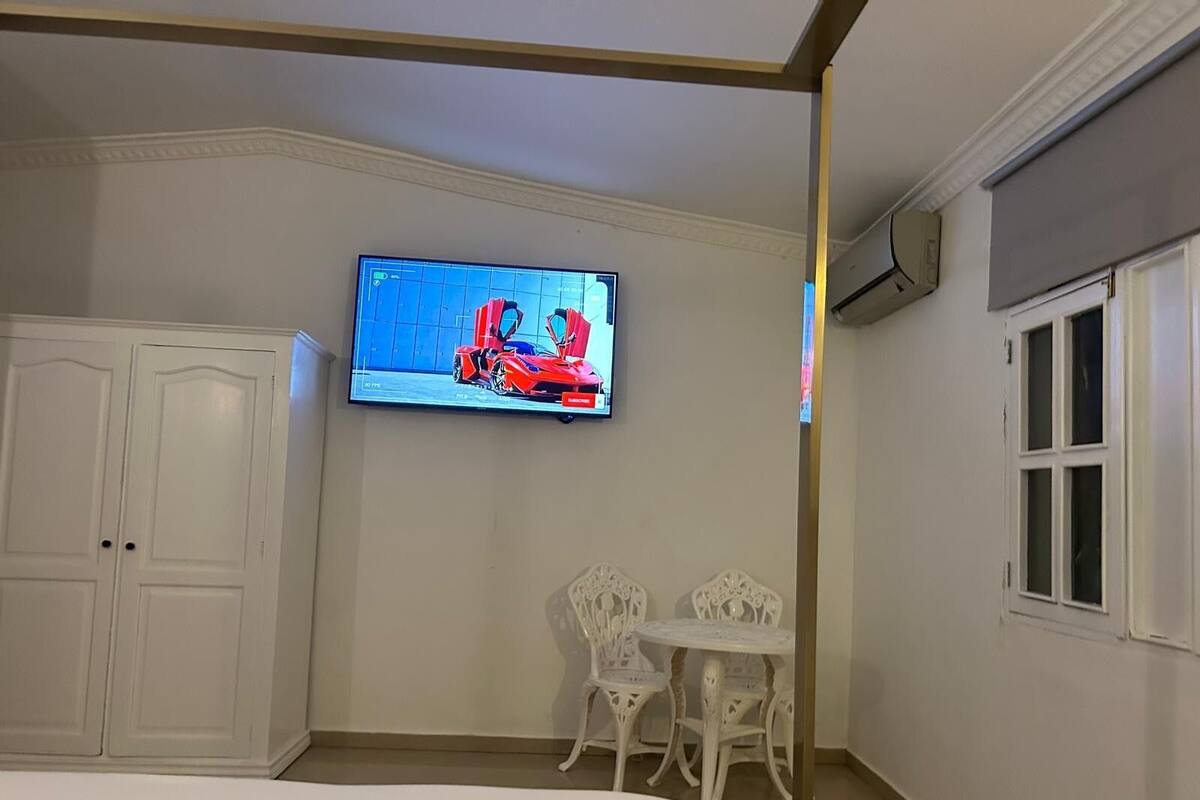 Ein Hotelzimmer mit Bett, einem an der Wand montierten Fernseher, der einen Sportwagen zeigt, einem weißen Kleiderschrank und einem kleinen weißen Tisch mit Stühlen.