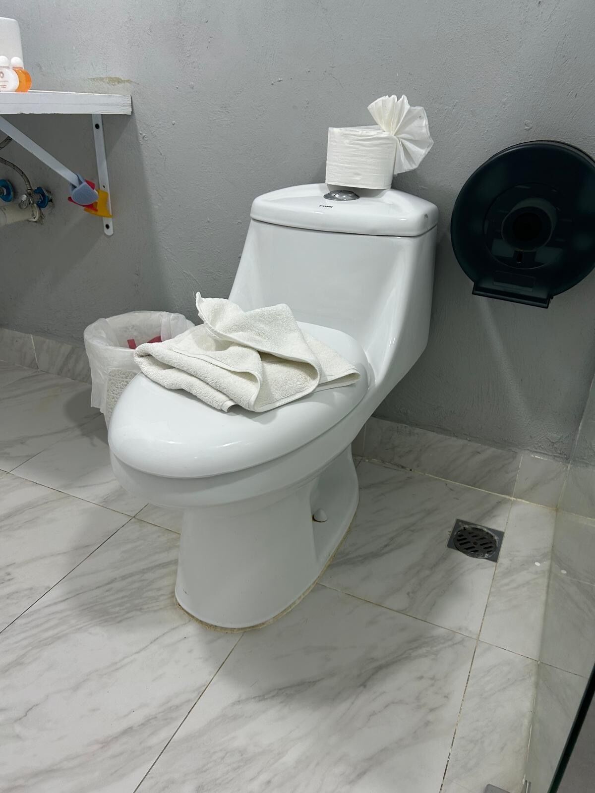 Un inodoro blanco con una caja de pañuelos y una toalla encima, un bote de basura y un dispensador de papel higiénico negro en la pared.