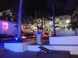 Un espace extérieur nocturne avec un banc blanc, des palmiers et un bâtiment dont les fenêtres sont illuminées.