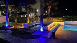 Un espace extérieur avec une piscine, agrémenté d’une fontaine illuminée de lumière bleue, d’un coin salon et de palmiers.