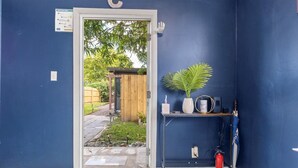 Ein Zimmer mit blauer Wand, einer Holztür, die in einen Garten führt, einem kleinen Tisch mit Pflanze, einer Vase und einem Feuerlöscher.
